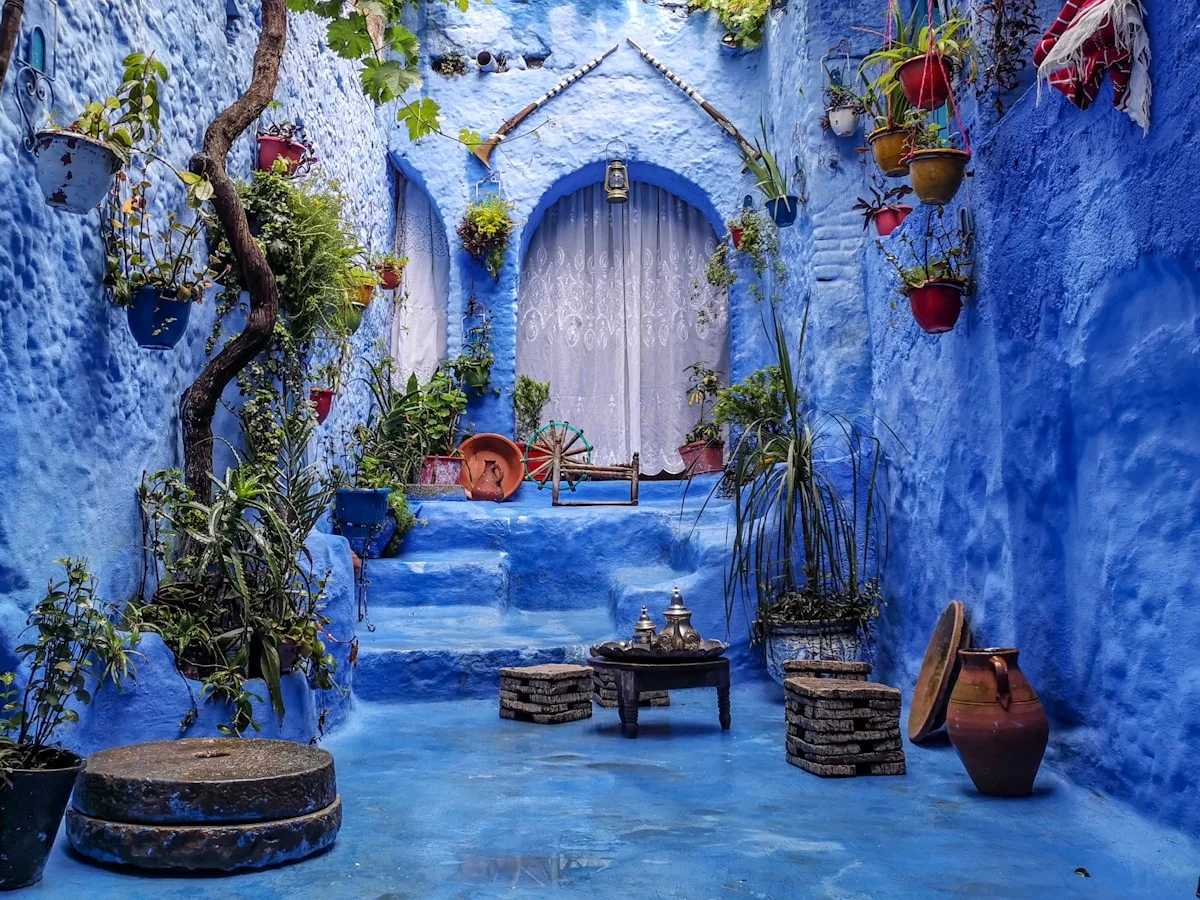Chefchaouen, Morocco — the iconic blue-washed medina buildings with colorful flower pots, narrow winding streets, and distant Rif Mountains under clear sky